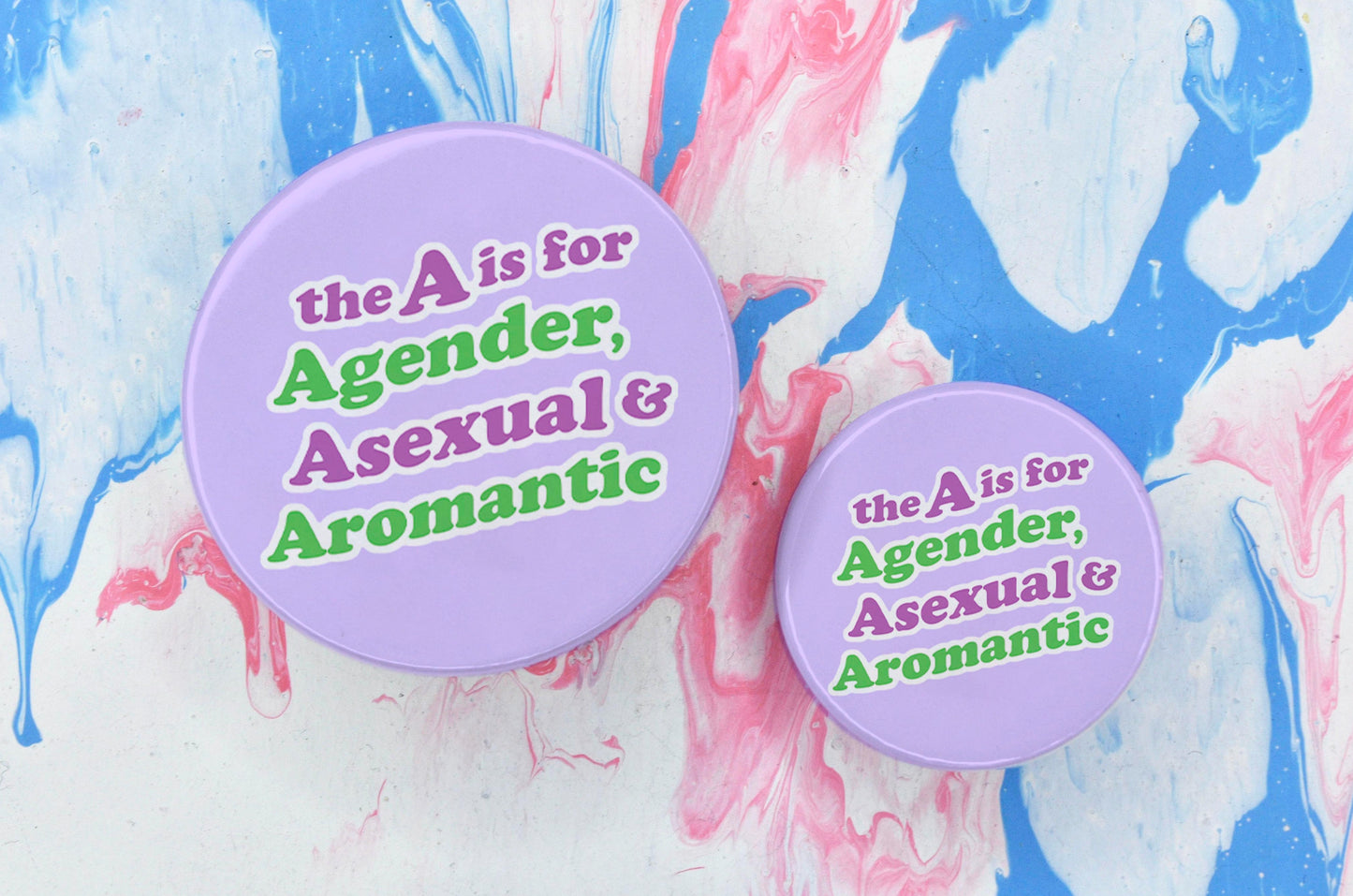 Two badges in sizes medium and small respectively, each featuring a pastel purple background with text in a bubble-like font reading "The A is for Agender, Asexual and Aromantic". The text is split across four lines, with the text of the first line being dark pink, the second being green, the third dark pink and the fourth green - this is to represent the colours within the agender, ace and aro flags.