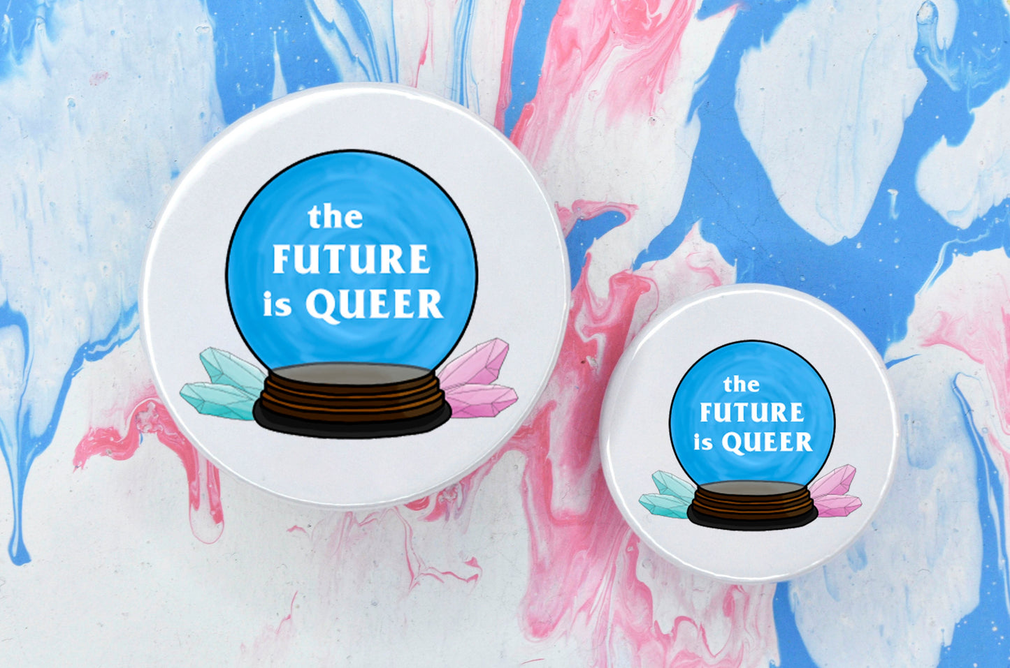 Two pin badges in sizes medium and small, both featuring an illustration of a blue crystal ball with white text reading "the future is queer". There are two blue and two pink crystal on the left and right of the crystal ball respectively, and the badge has a white background.