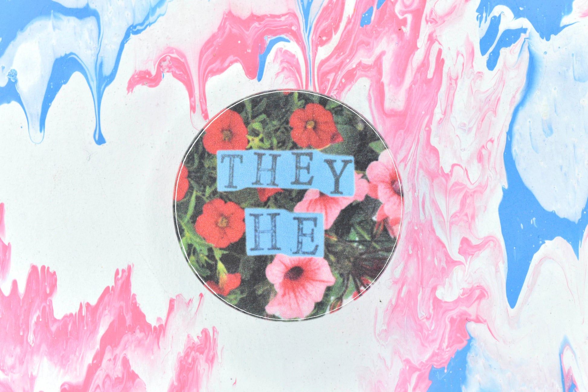 A pin badge featuring a floral background showing some red and dark pink flowers surrounded by green leaves. Overlaid are hand cut pieces of blue paper with hand stamped uppercase black lettering reading "they" and "he".