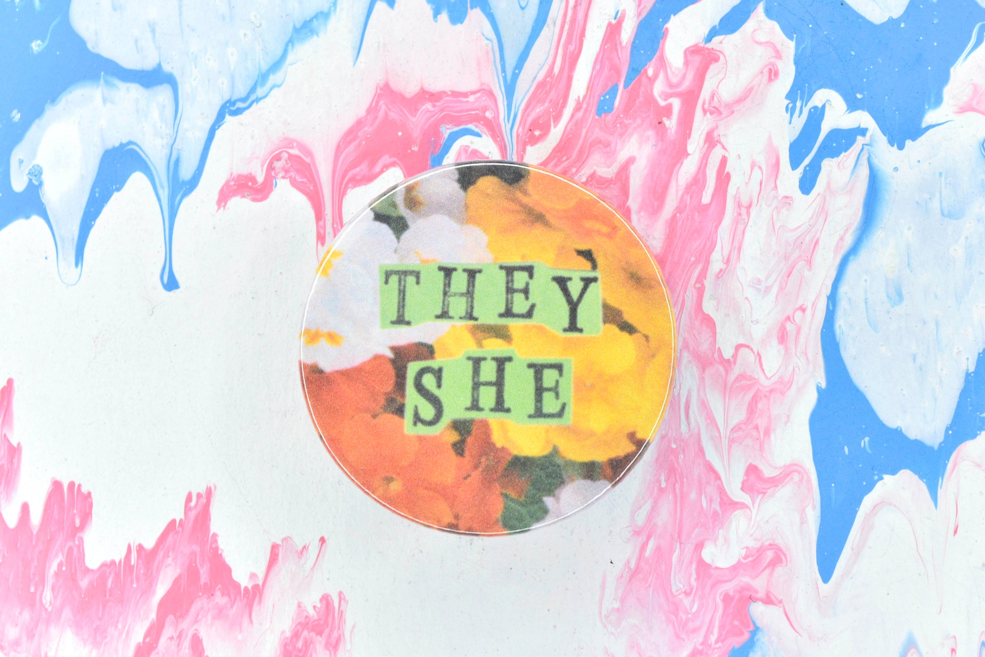 A pin badge featuring a floral background showing some yellow, white and orange primroses. Overlaid are hand cut pieces of green paper with hand stamped uppercase black lettering reading "they" and "she".