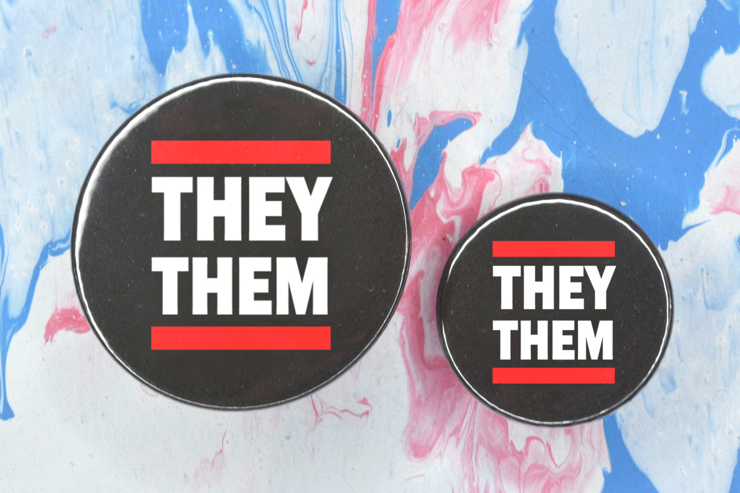 Two circular pin badges in sizes medium and small each with a black background, two rows of bold white upper case text and a solid red line above and below the text. The top row reads "they" and the bottom row reads "them".