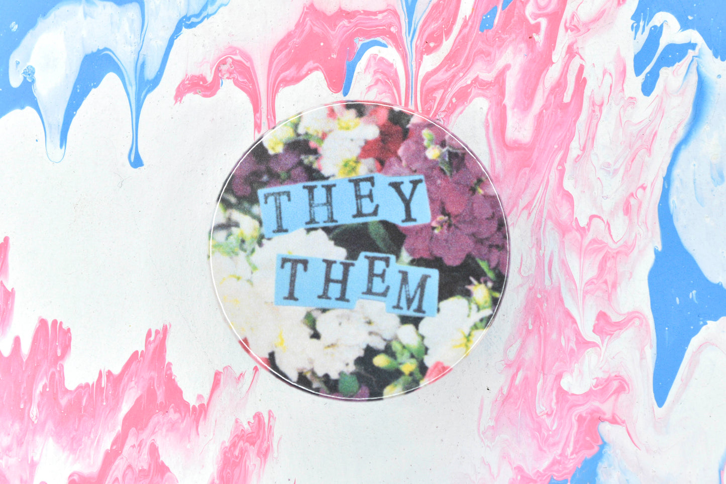 A pin badge featuring a floral background showing some pink and white flowers. Overlaid are hand cut pieces of pastel blue paper with hand stamped uppercase black lettering reading "they" and "them".