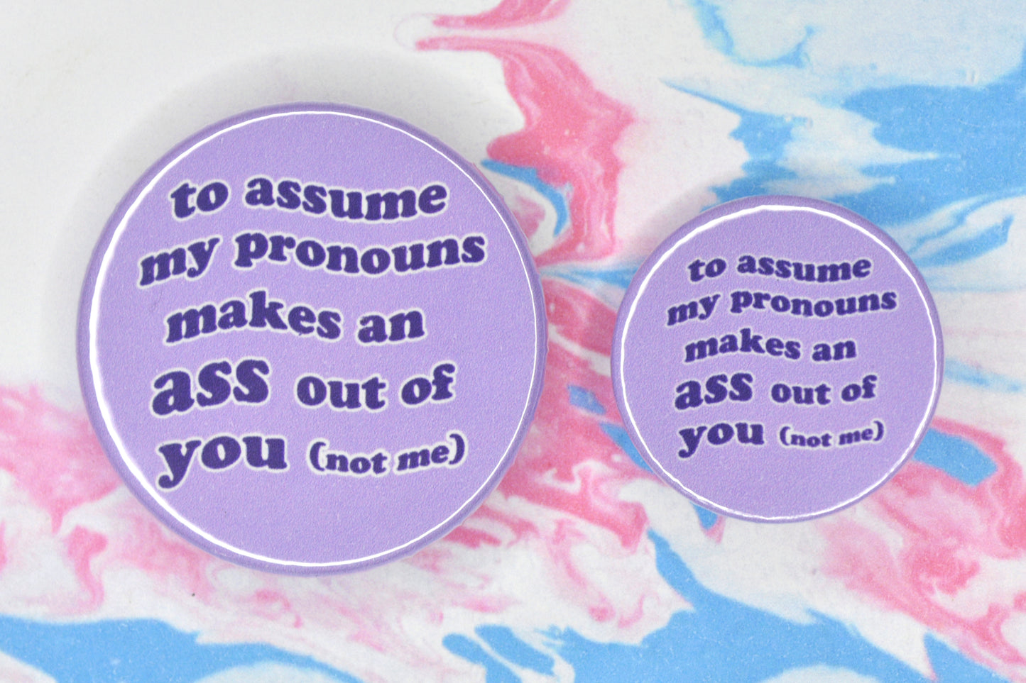 Two badges in sizes medium and small respectively, each featuring a light purple background with wavy bubble-like text in a darker purple text reading "to assume my pronouns makes an ass out of you (not me)".