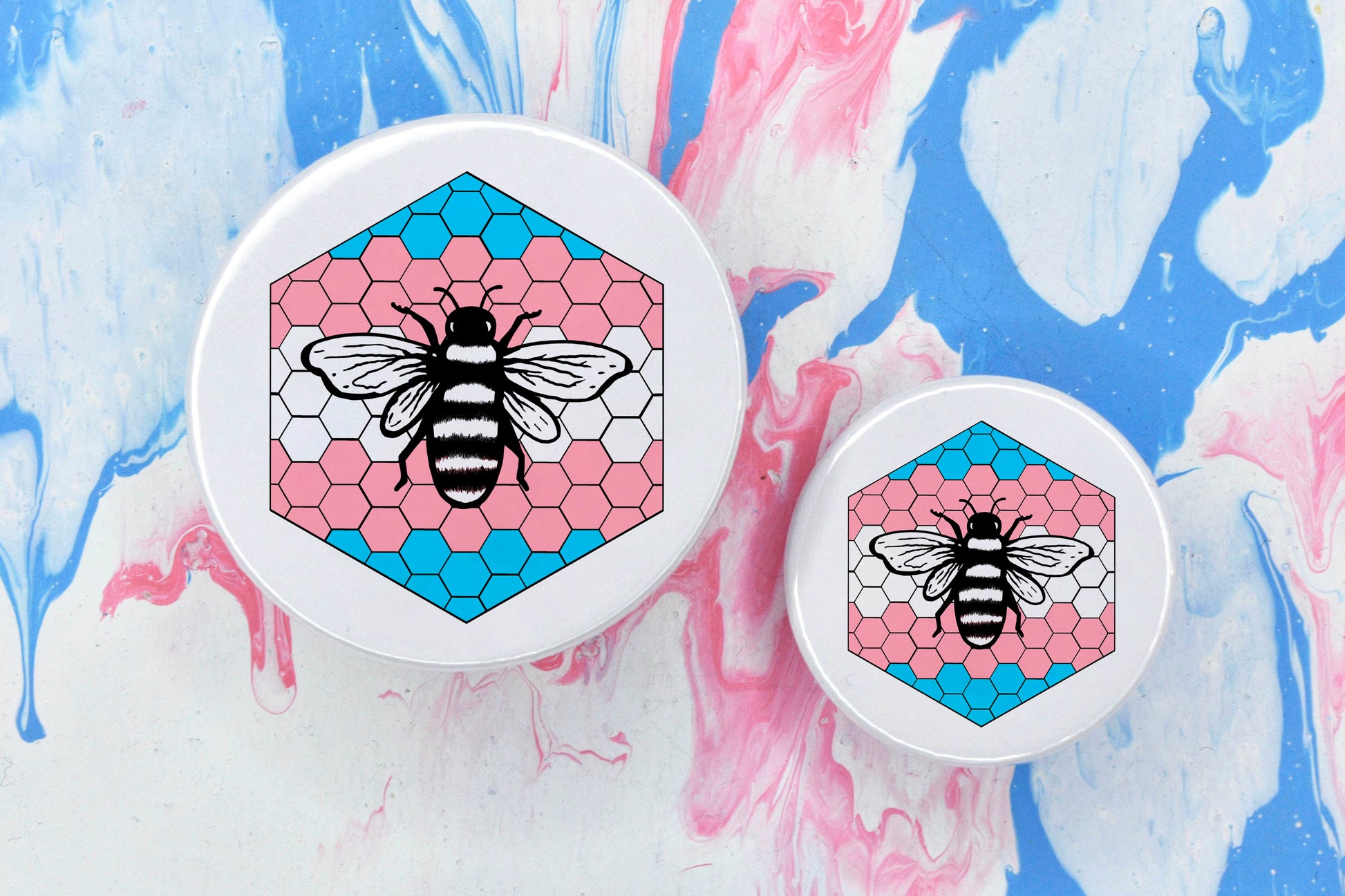 Two badges in sizes medium and small respectively, each featuring a white background with a black and white illustration of a bee overlaid on a bee hive pattern which is coloured in with the trans pride flag colours (blue, pink and white).