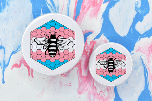 Two badges in sizes medium and small respectively, each featuring a white background with a black and white illustration of a bee overlaid on a bee hive pattern which is coloured in with the trans pride flag colours (blue, pink and white).