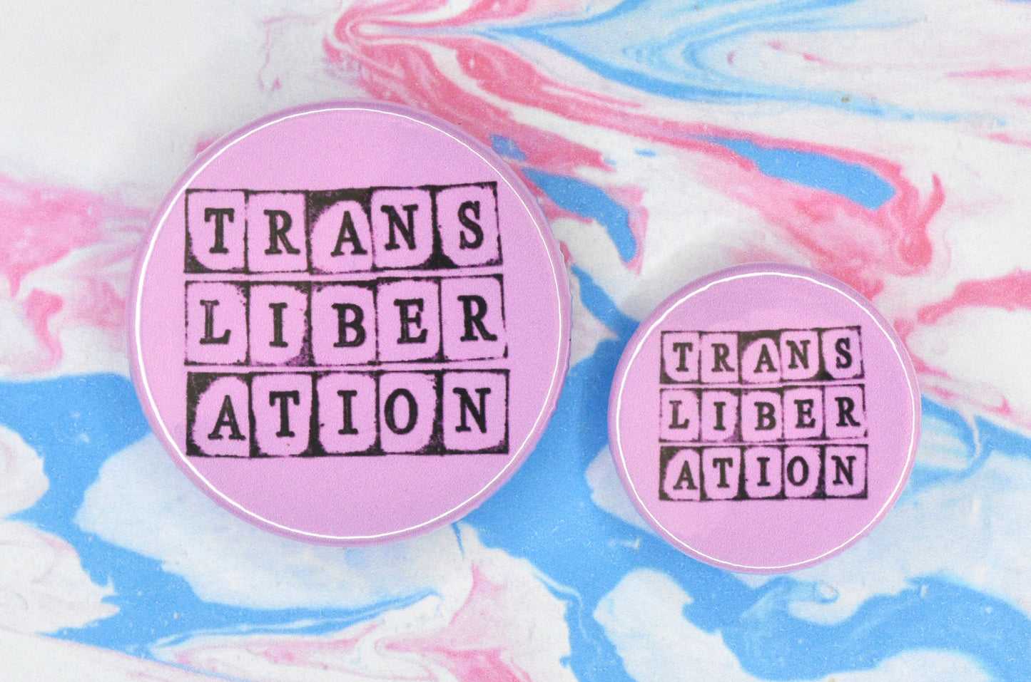 Two badges in sizes medium and small respectively, each featuring a pink background with text in a hand-stamped font reading "trans liberation".