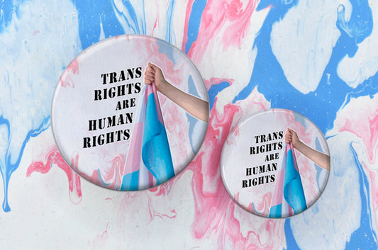 Two badges in sizes medium and small respectively, each featuring a black stencil-like font reading "trans rights are human rights" next to a photo of a hand holding up a trans flag. The background of the badges is a pink, white and blue paint pouring design.