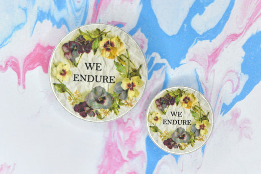 Two badges in sizes medium and small respectively, each featuring a collage design consisting of a vintage photo of some flowers forming a circular border with black uppercase serif text in the centre reading "We endure". The background is an off-white crumpled paper texture.