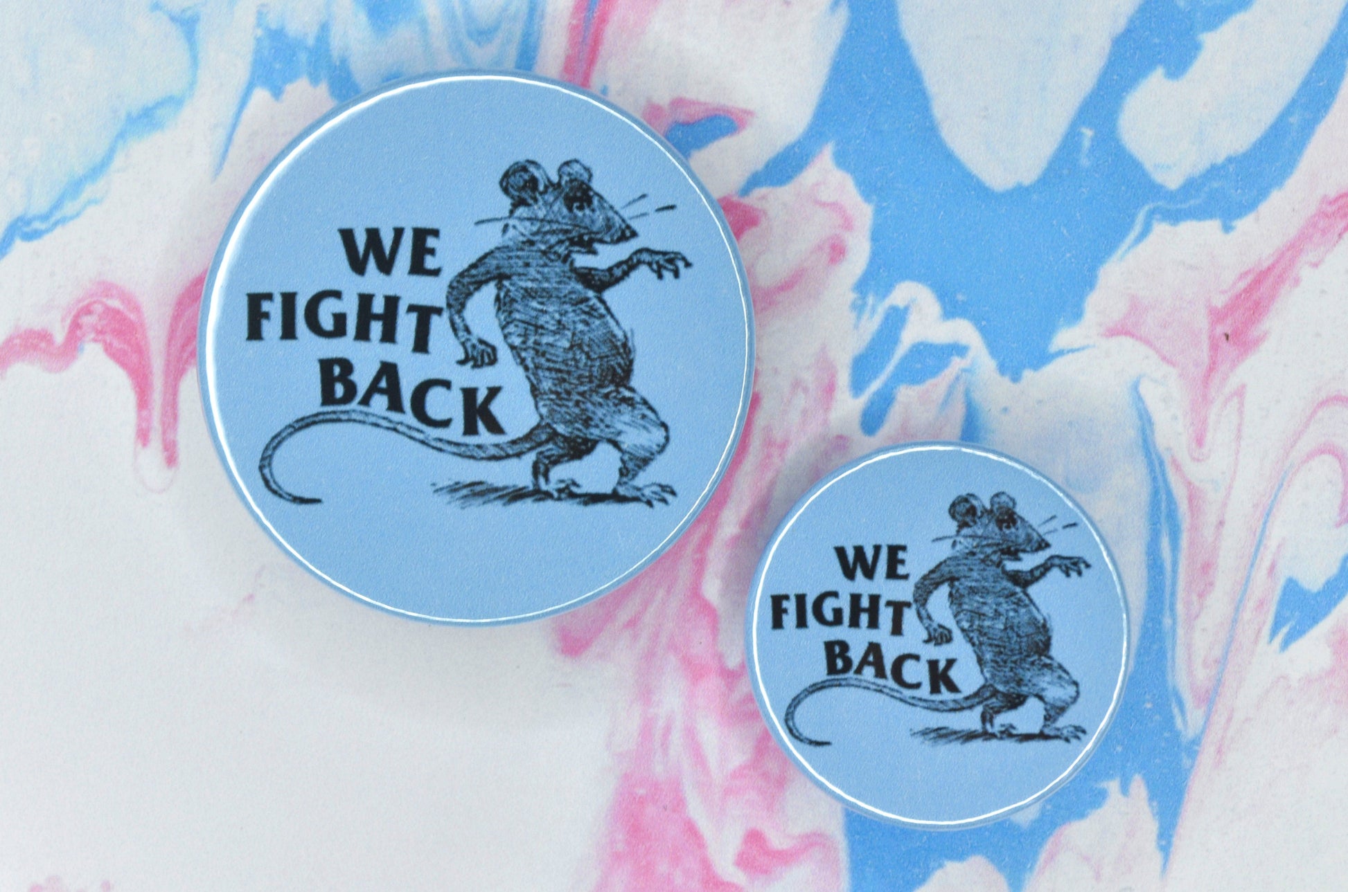 Two badges in sizes medium and small respectively, each featuring a blue background with a vintage illustration of a rat standing on its hind legs with one of its front paws outstretched. There is large black uppercase text to the left of the rat reading "we fight back".