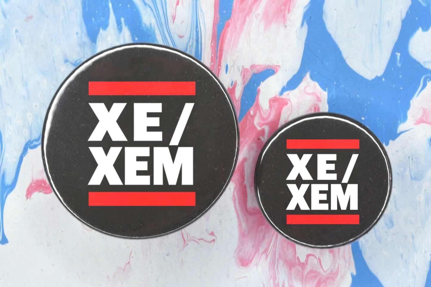 Two circular pin badges in sizes medium and small each with a black background, two rows of bold white upper case text and a solid red line above and below the text. The top row reads "xe" with a forward slash and the bottom row reads "xem".