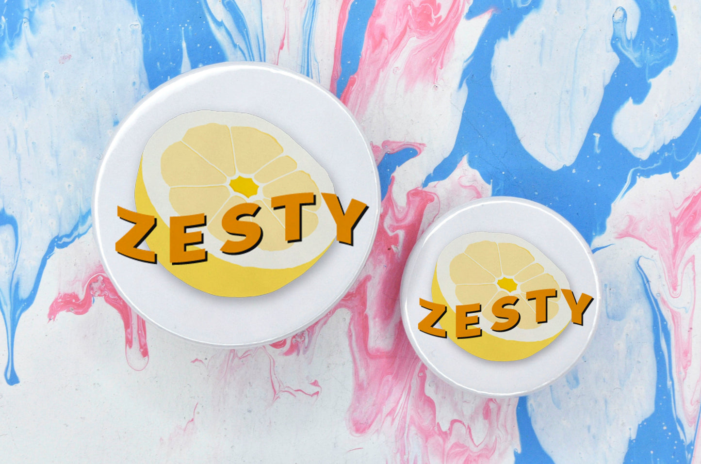 Two circular pin badges in sizes medium and small, each with a white background and an illustration of a yellow lemon in the centre. There is orange uppercase text across the centre of the lemon reading "zesty".