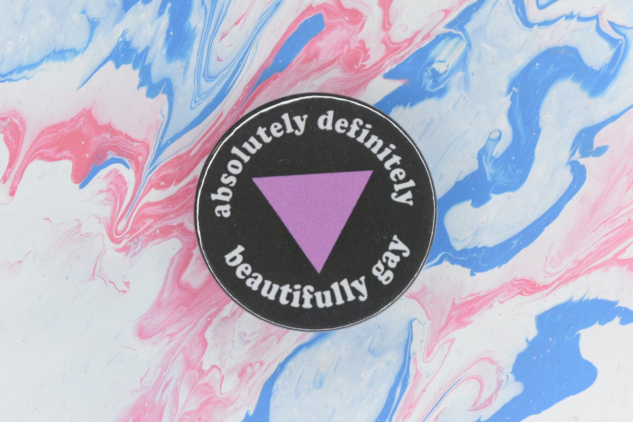 A single pin badge in size medium featuring a black background with a pink triangle in the centre and white text around the outside reading "absolutely definitely beautifully gay".